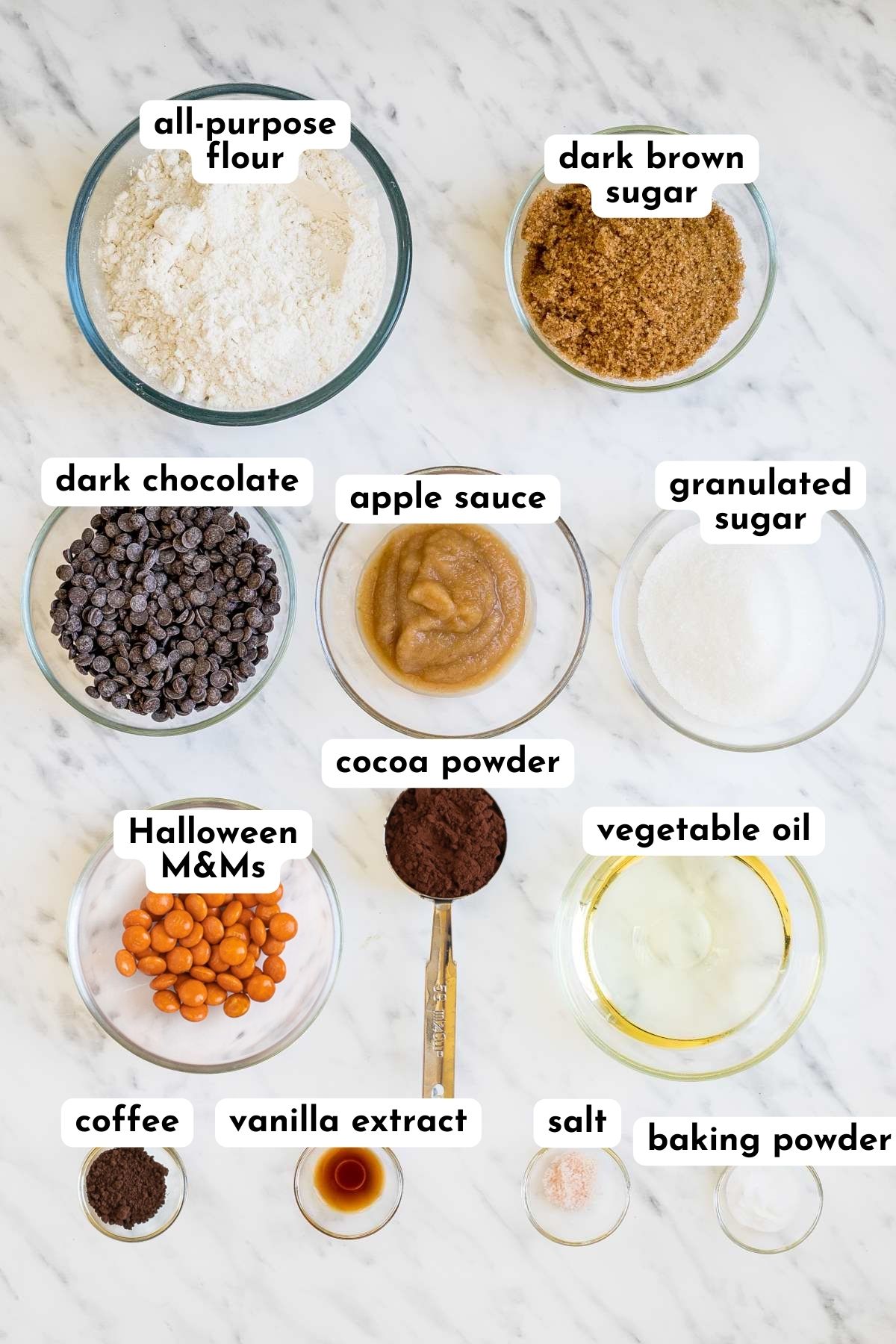 ingredients of halloween cookies in small glass bowls like flour, sugar, chocolate chips, orange candies, cocoa powder, apple sauce, oil, salt, baking soda, and coffee