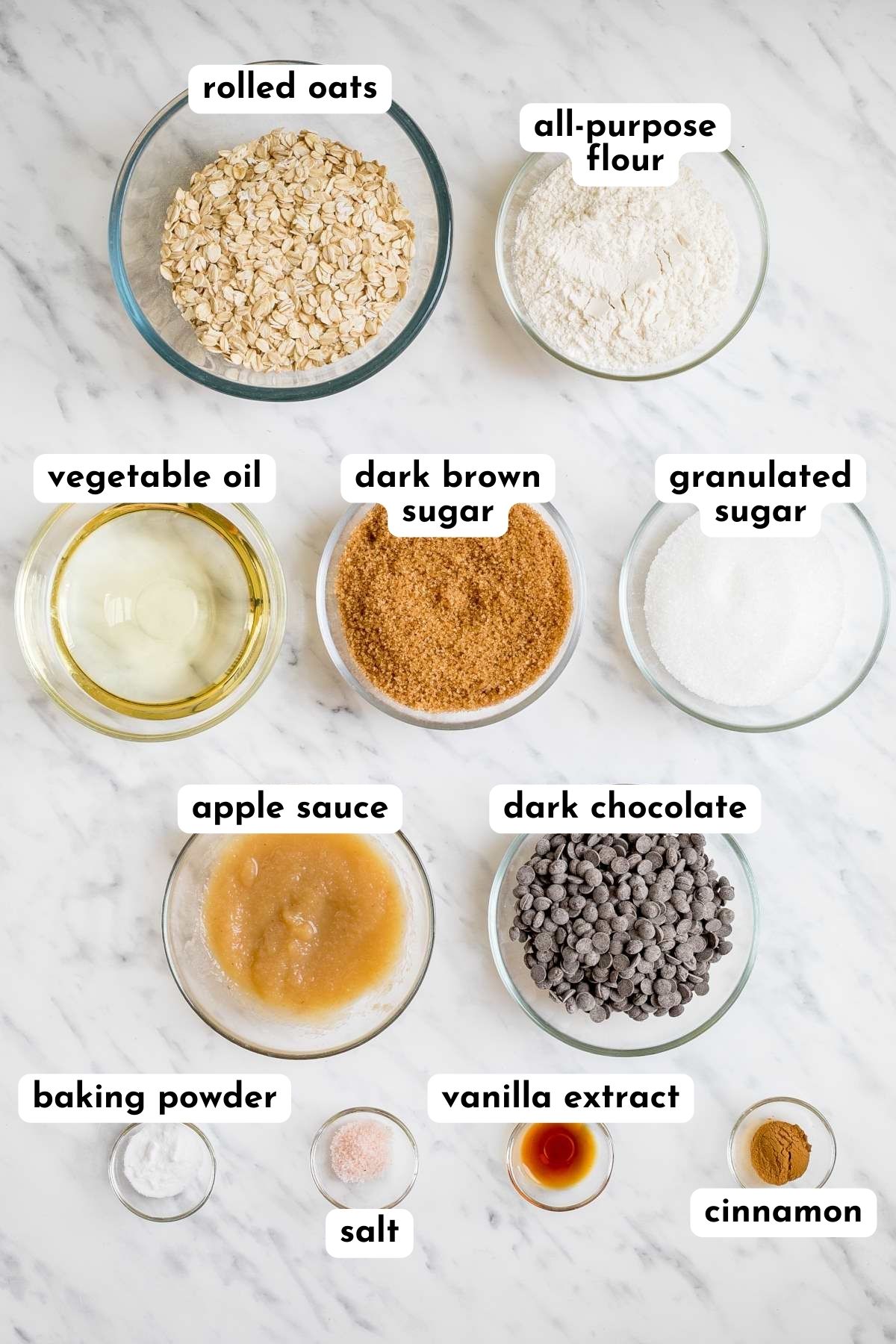Ingredients of oatmeal chocolate chip cookies in small glass bowls like flour, rolled oats, sugar, dark chocolate chips, apple sauce, oil, salt, cinnamon, vanilla extract, and baking soda.