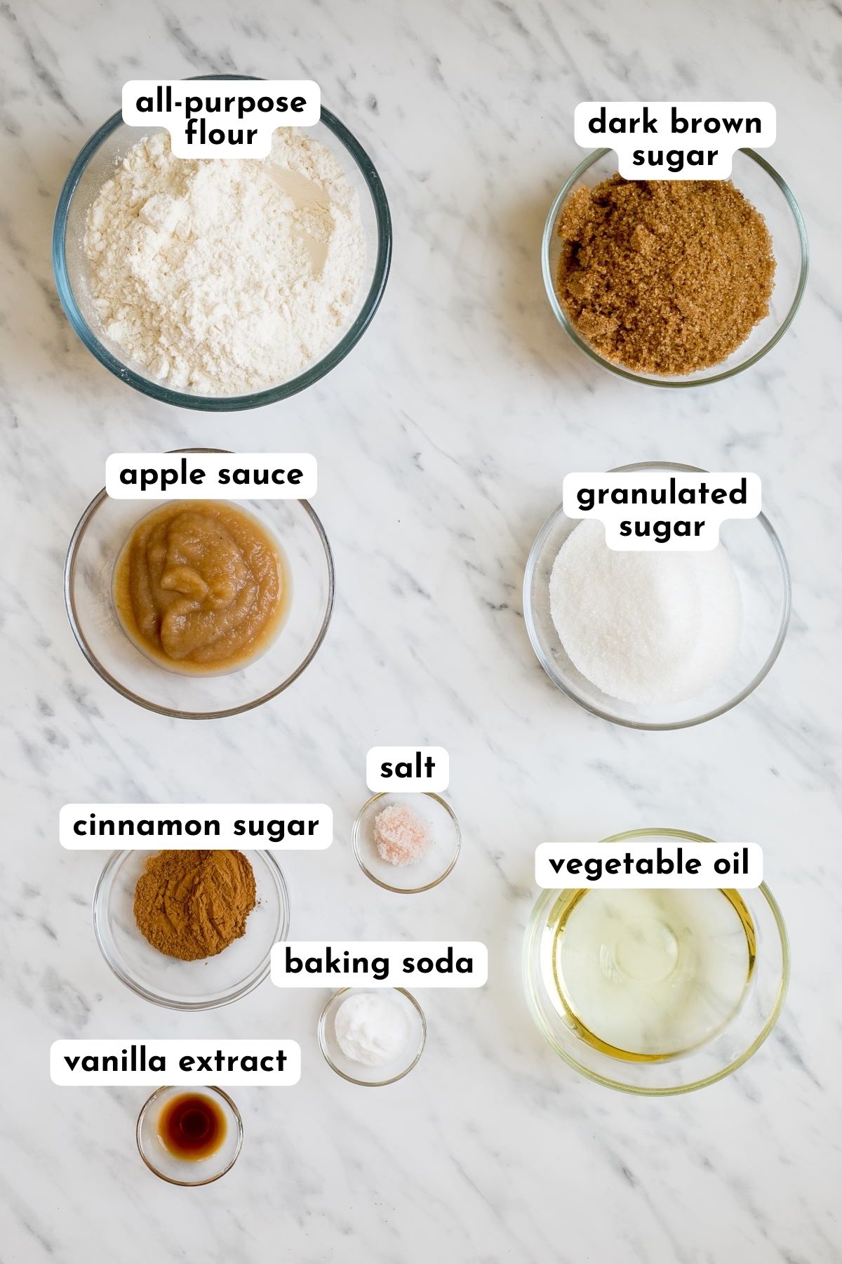 The ingredients of snickerdoodle cookies in small glass bowls like flour, brown sugar, white sugar, oil, apple sauce, cinnamon, vanilla extract, salt, and baking soda. 