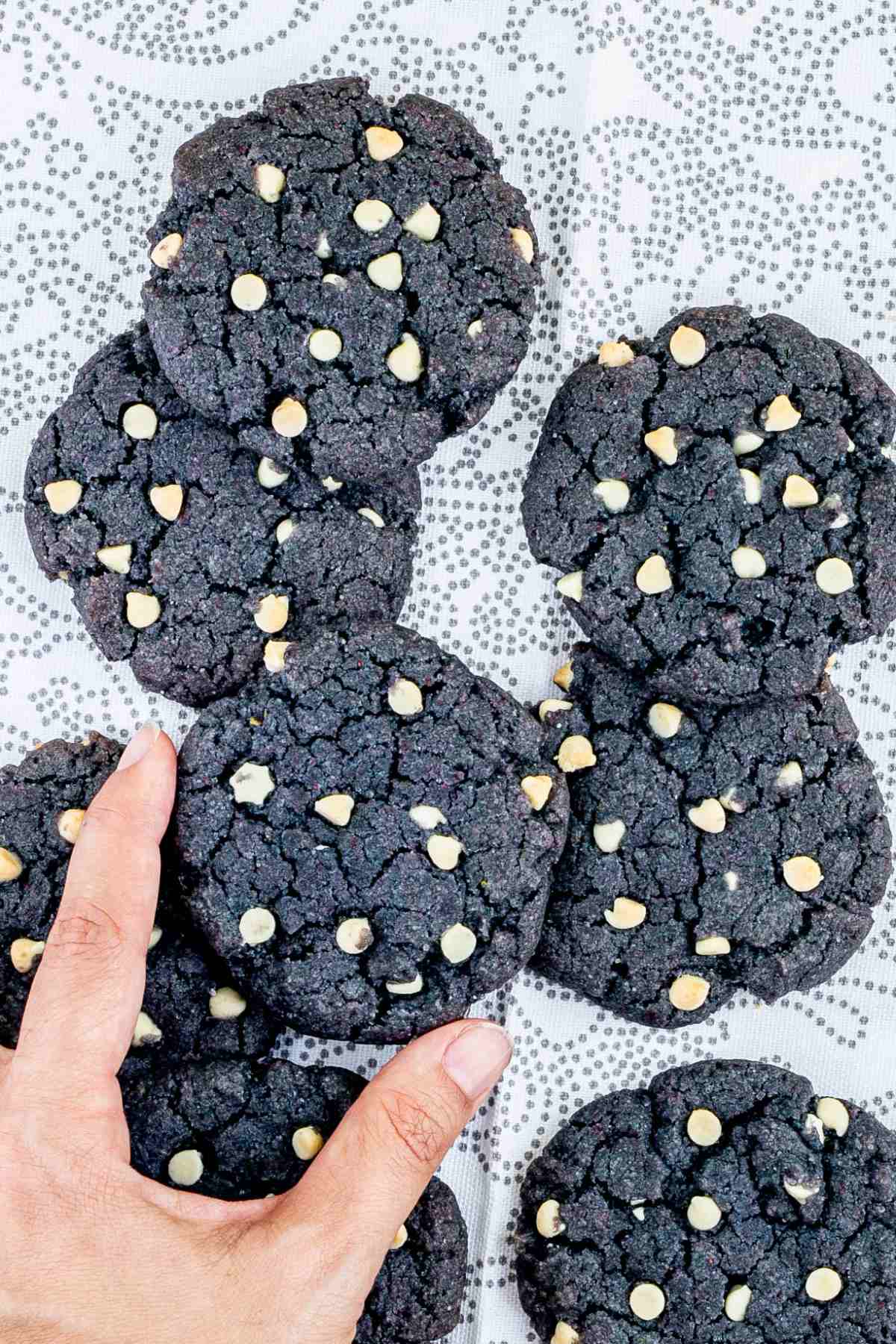 Blue-purple cookies with white chocolate chips on a white-grey surface. A hand is taking one.