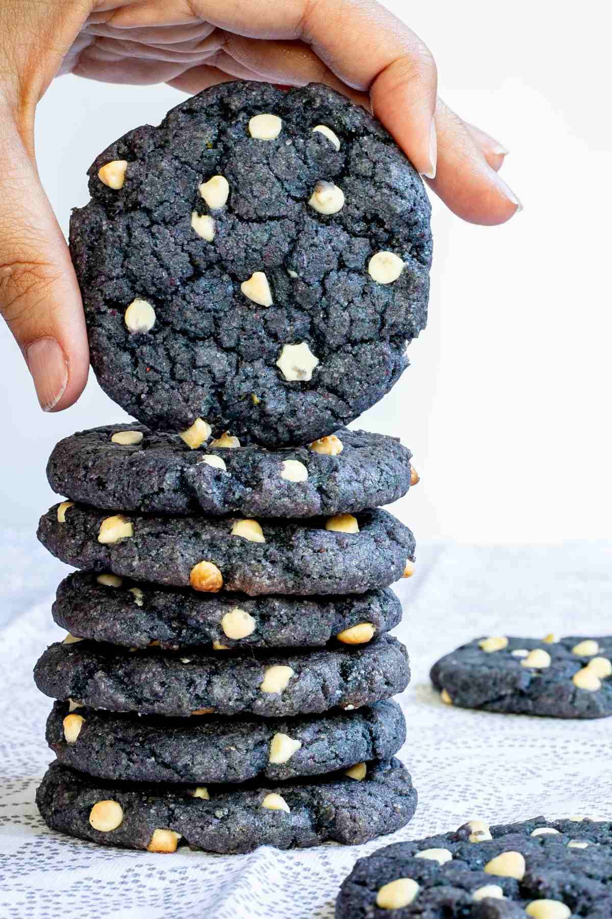 A stack of blue-purple cookies with white chocolate chips on a white-grey surface. A hand is taking one from the top.