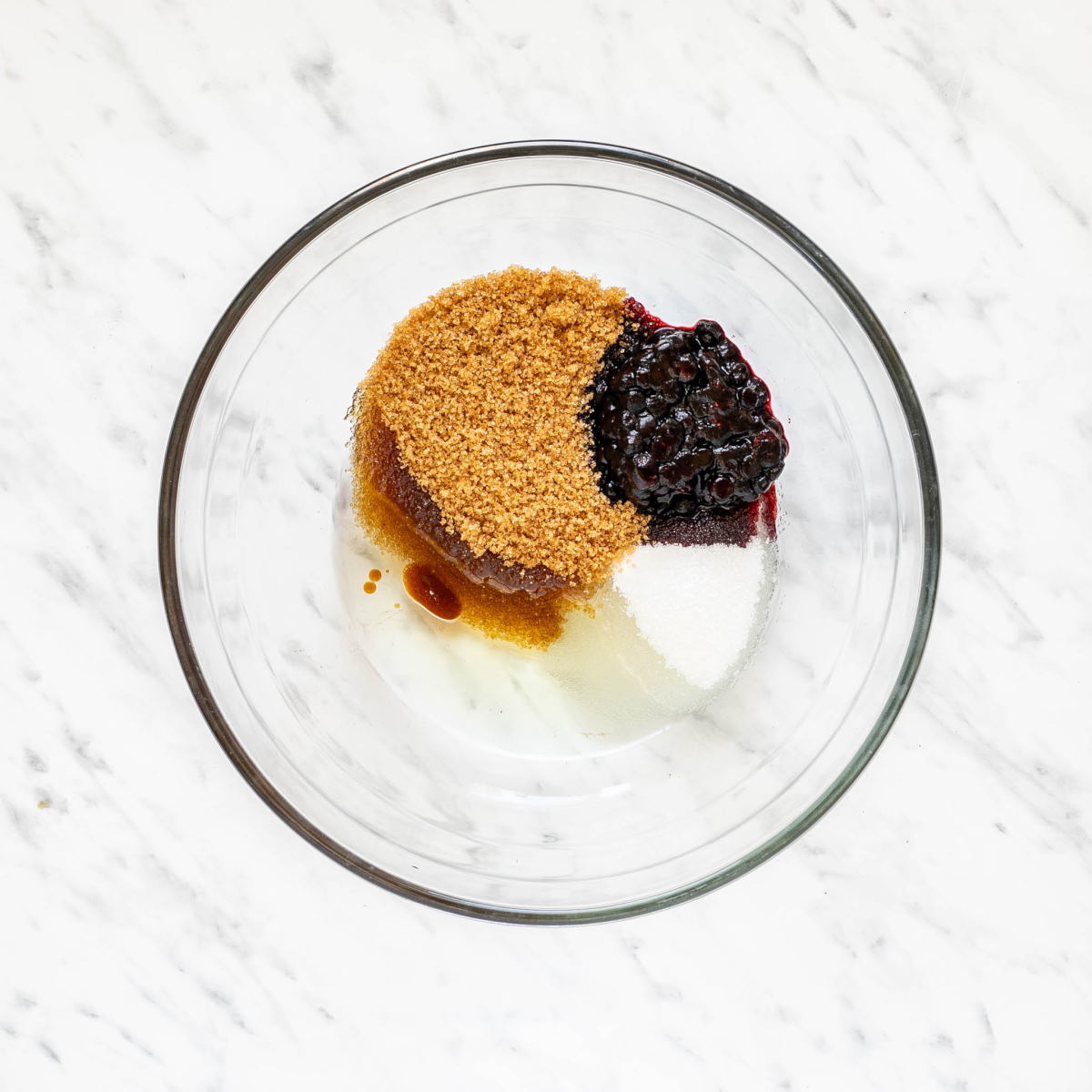 A glass bowl with soft small blueberries, white and brown sugar, and oil.