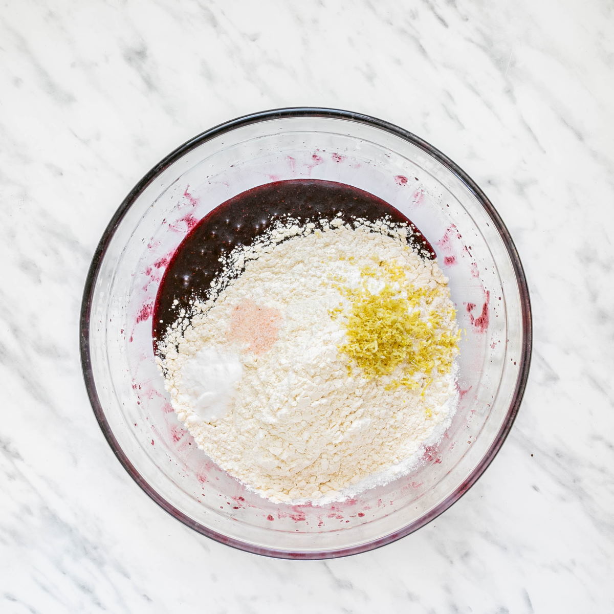 A glass bowl with dark purple liquid and a heap of flour, lemon zest, salt, and white powder.