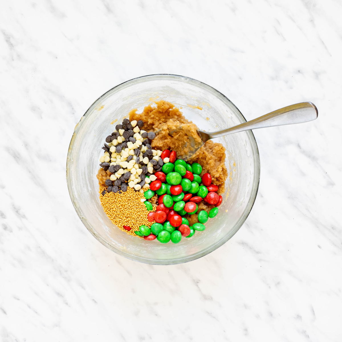 A glass bowl with light brown cookie down with red and green m&ms, chocolate chips, tiny white sugar snowflakes, and tiny green pine trees.