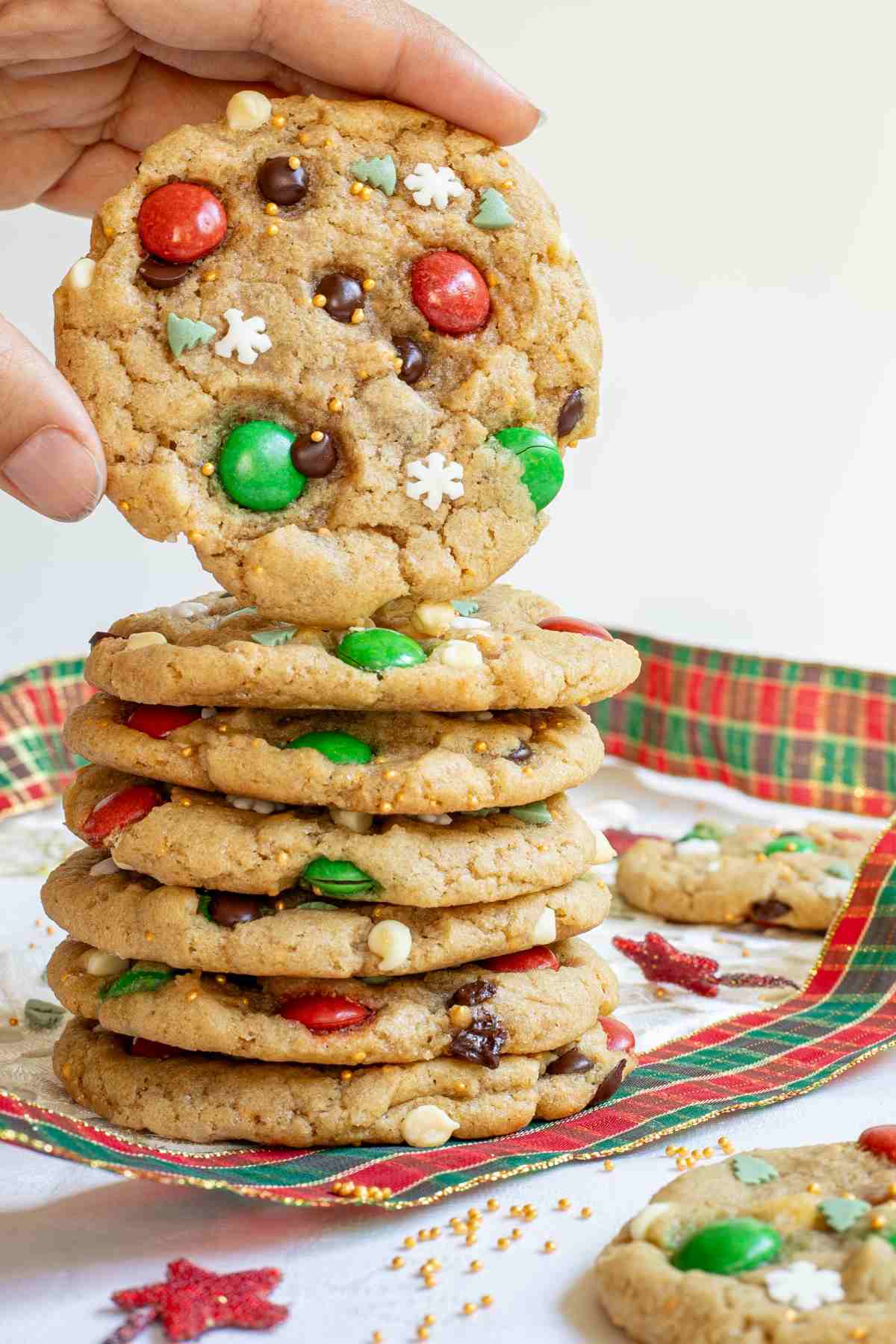 A stack of light brown cookies with red and green m&ms, chocolate chips, tiny white sugar snowflakes, and tiny green pine trees. A hand is taking one from the top.