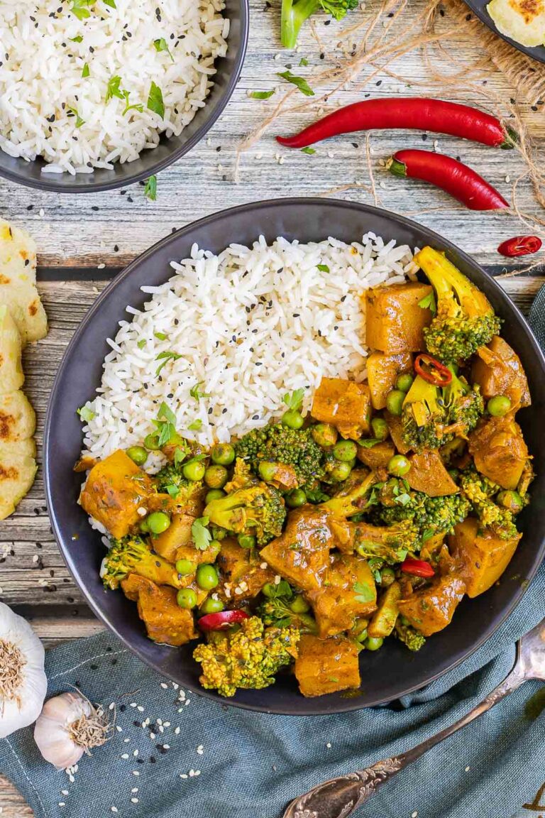 Black plate with rice topped with diced potatoes, broccoli, and green peas glazed in an orange sauce. More rice and flatbreads are placed next to it.