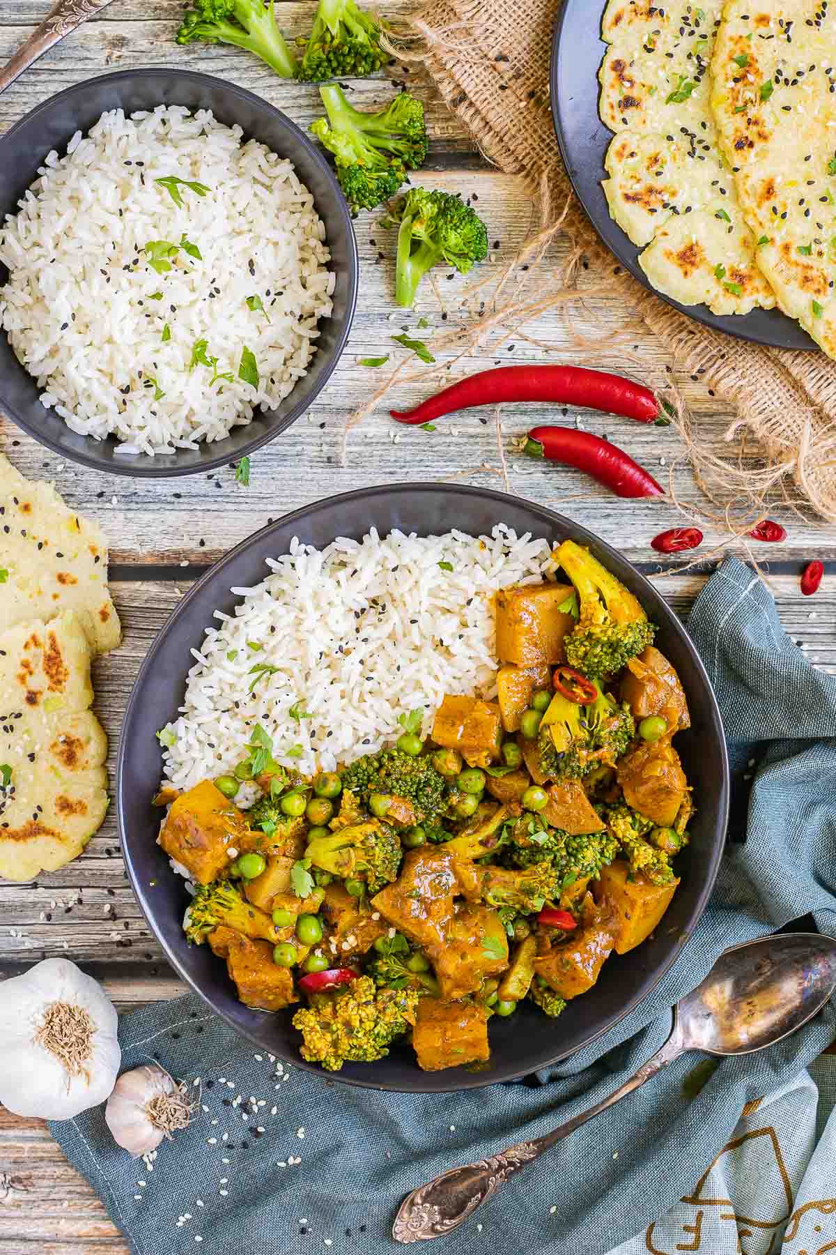 Black plate with rice topped with diced potatoes, broccoli, and green peas glazed in an orange sauce. More rice and flatbreads are placed next to it.