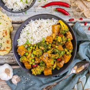 Black plate with rice topped with diced potatoes, broccoli, and green peas glazed in an orange sauce. More rice and flatbreads are placed next to it.