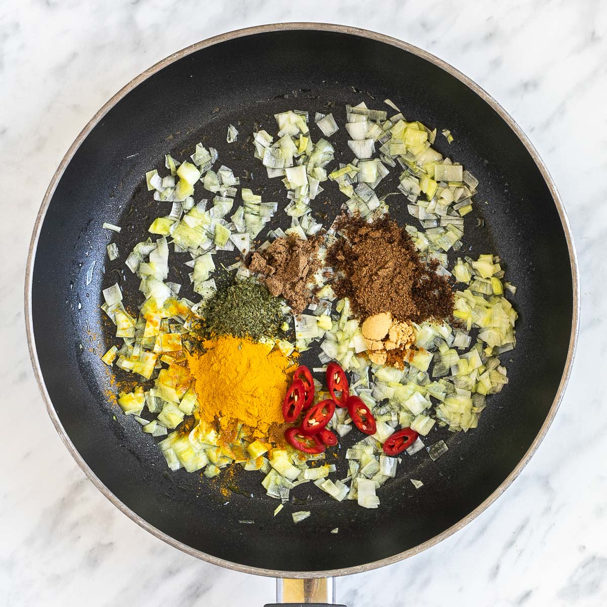 Black frying pan with finely chopped onion and small heaps of colorful herbs and spices.