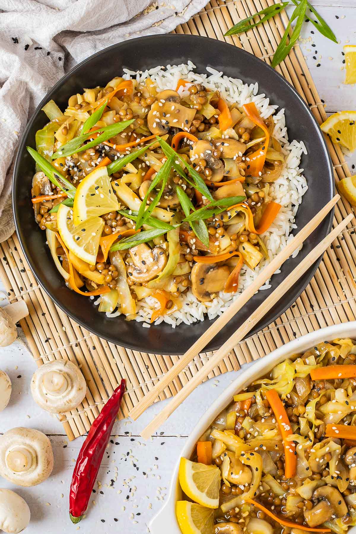 Black plate with white rice topped with stir fired cabbage, mushroom, carrot and lentils. A pair of chopsticks is placed on the side. More stir fried veggies are placed in a separate white bowl.
