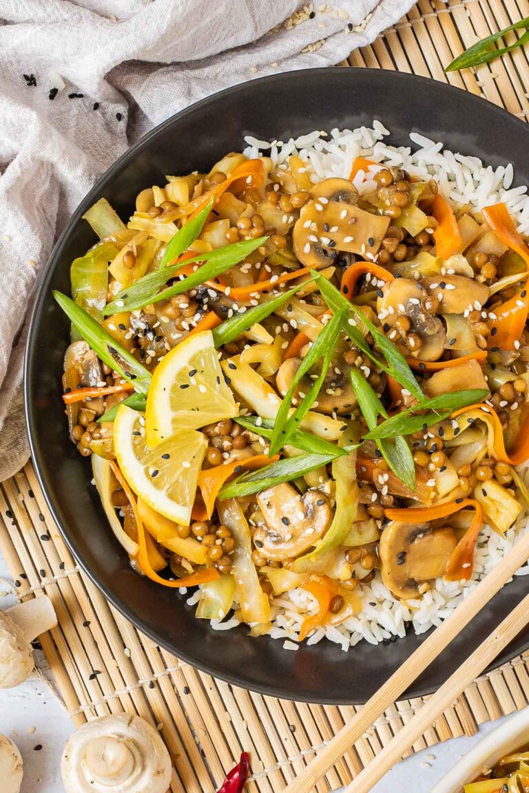 Black plate with white rice topped with stir fired cabbage, mushroom, carrot and lentils. A pair of chopsticks is placed on the side.