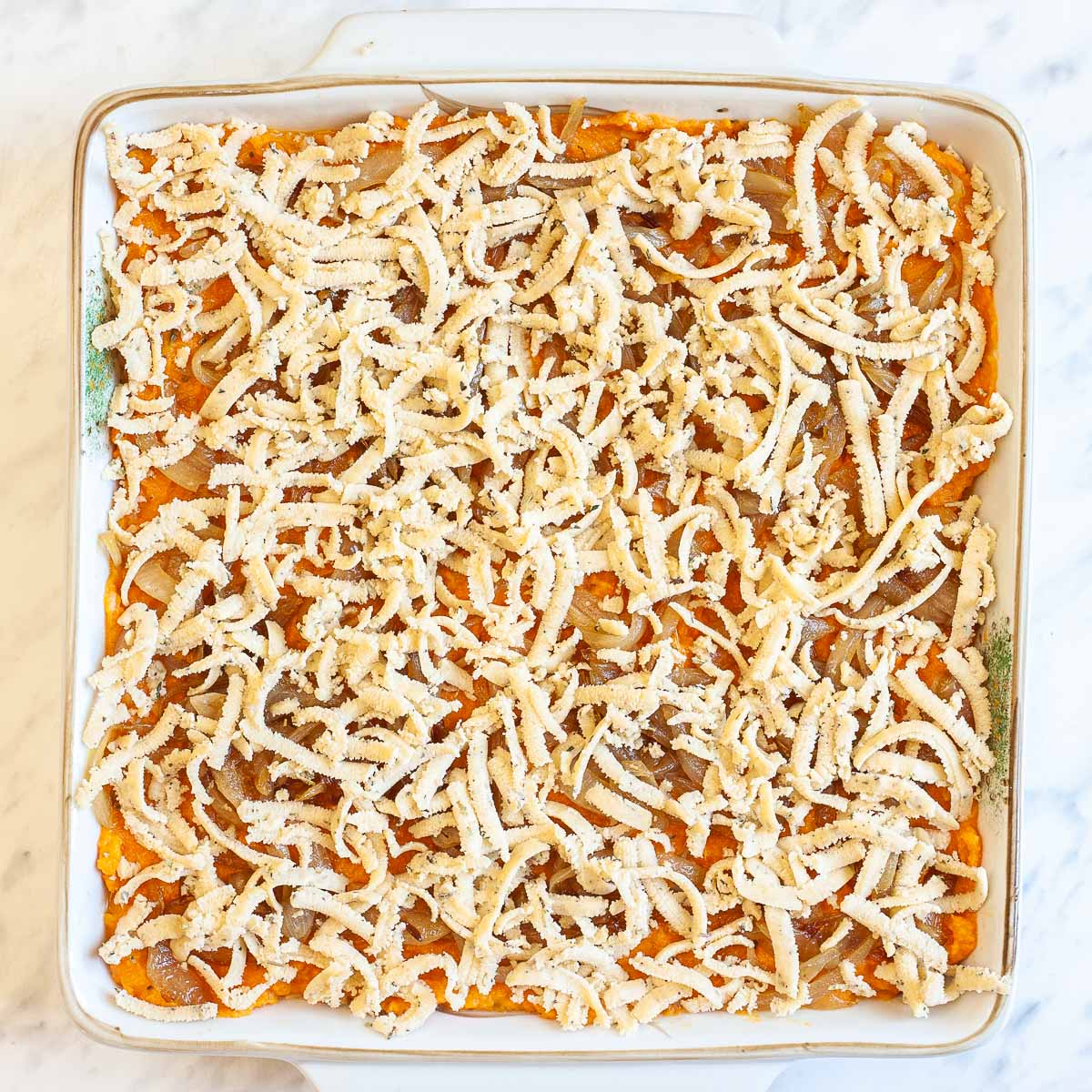A white 9x9 baking dish with creamy mashed orange sweet potatoes topped with light brown caramelized onion slices and grated dough.