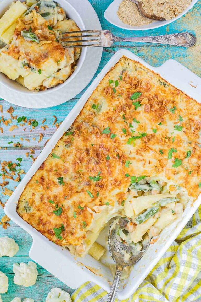 A baking dish with penne pasta in creamy sauce, chickpeas and green peas topped with breadcrumbs and freshly chopped herbs. The corner part is missing and served in a small plate on the side.