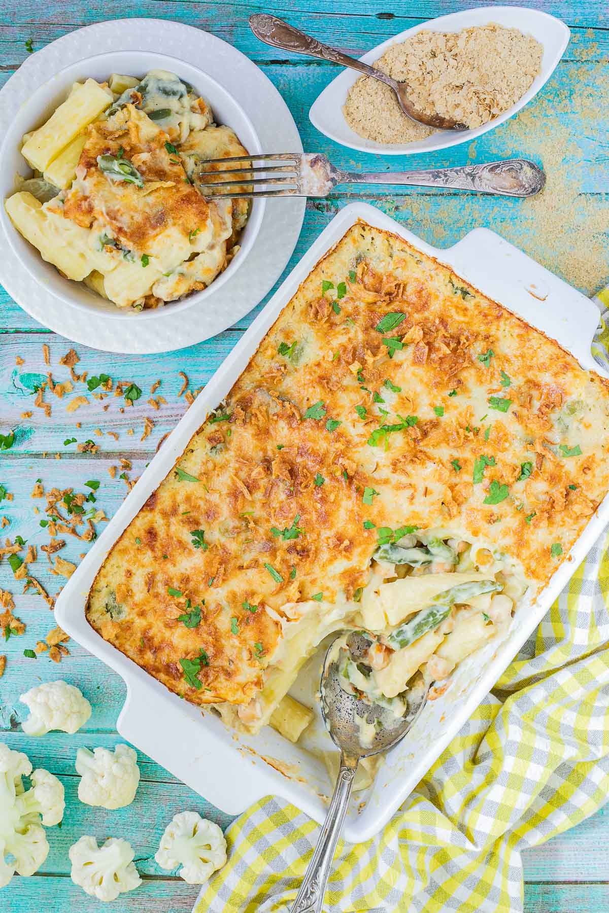 A baking dish with penne pasta in creamy sauce, chickpeas and green peas topped with breadcrumbs and freshly chopped herbs. The corner part is missing and served in a small plate on the side.
