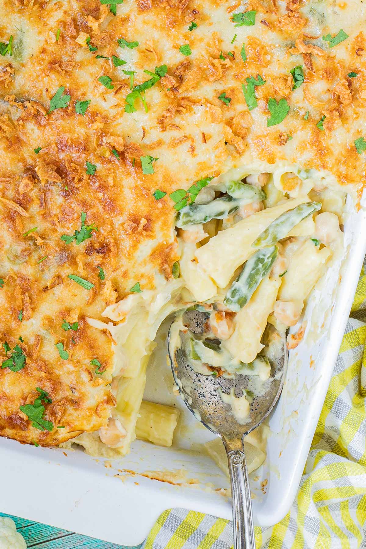 A baking dish with penne pasta in creamy sauce, chickpeas and green peas topped with breadcrumbs and freshly chopped herbs. The corner part is missing and served in a small plate on the side.