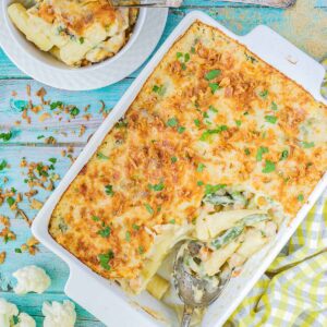 A baking dish with penne pasta in creamy sauce, chickpeas and green peas topped with breadcrumbs and freshly chopped herbs. The corner part is missing and served in a small plate on the side.