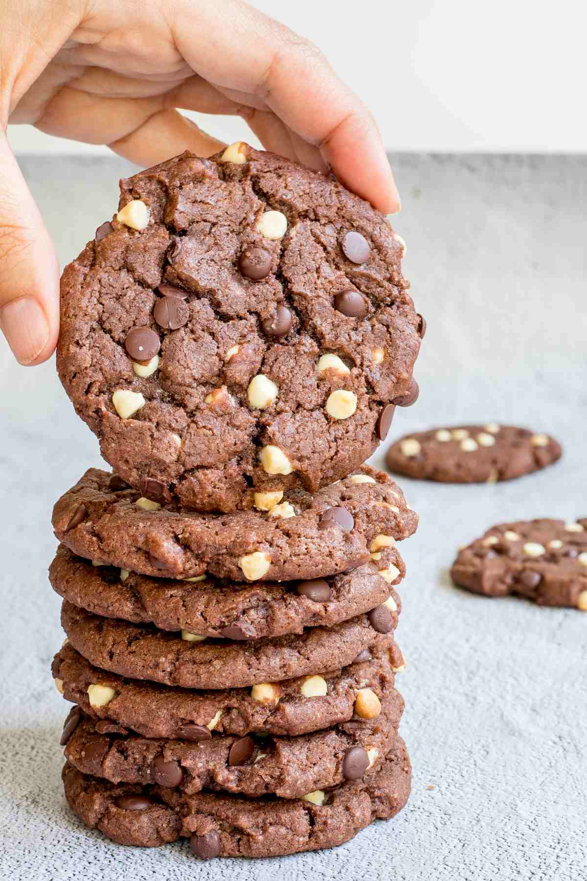 A stack of dark brown cookies with white and dark chocolate chips on a white surface. A hand is taking one from the top.