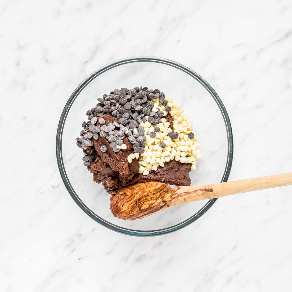 A glass bowl with uncooked dark brown cookie dough and lots of white and dark chocolate chips.