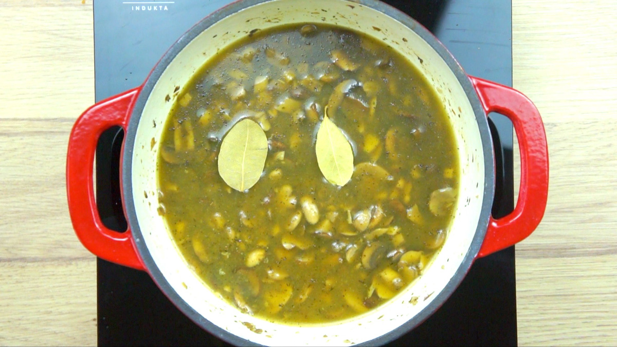 Red-white enameled Dutch oven with thin brown soup full of mushrooms and two bay leaves swimming on top.