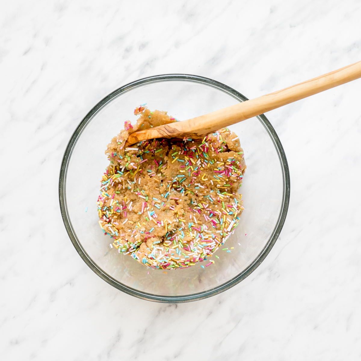A glass bowl with a wooden spatula and a light brown uncooked cookie dough with rainbow sprinkles.