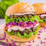 A burger bun with raffled green lettuce two lentil patties, purple coleslaw and light orange sauce.