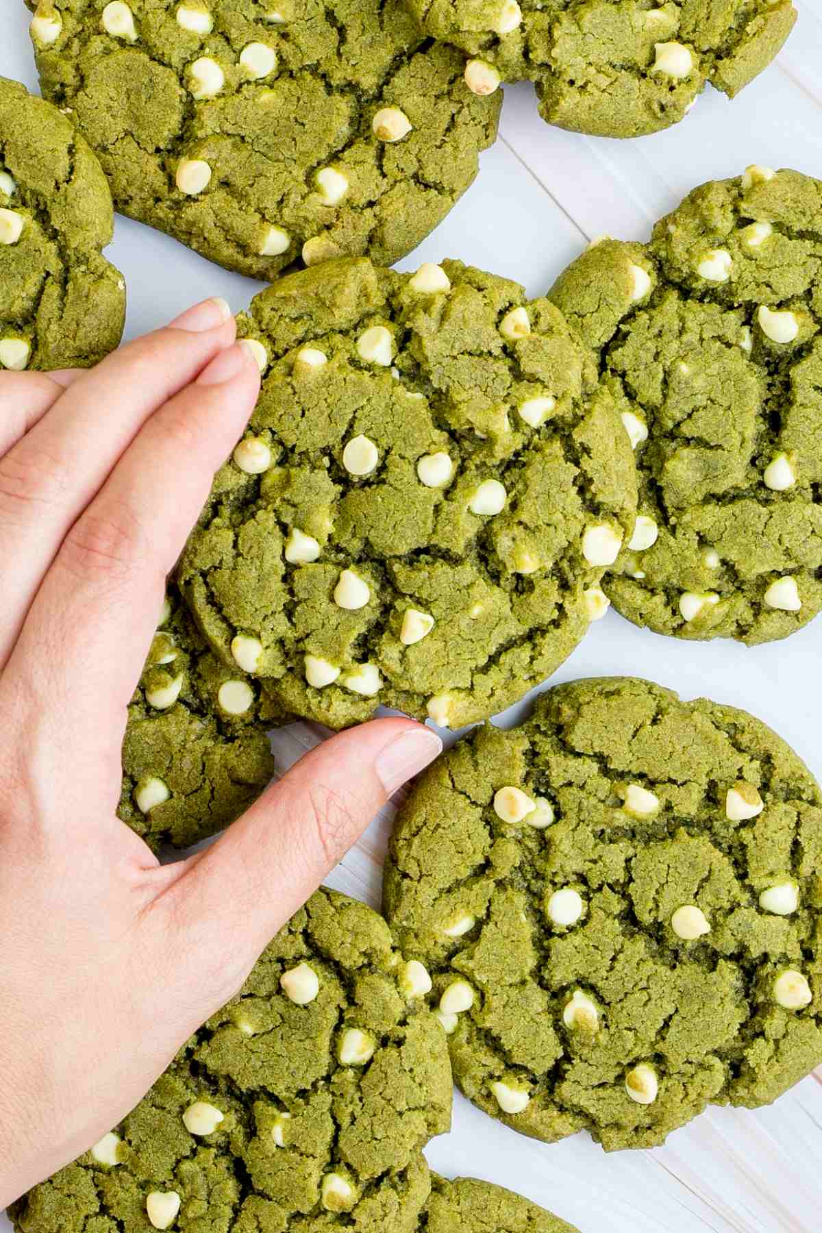 Lots of vibrant green cookies with white chocolate chips on a marble surface. A hand is taking one from the middle.