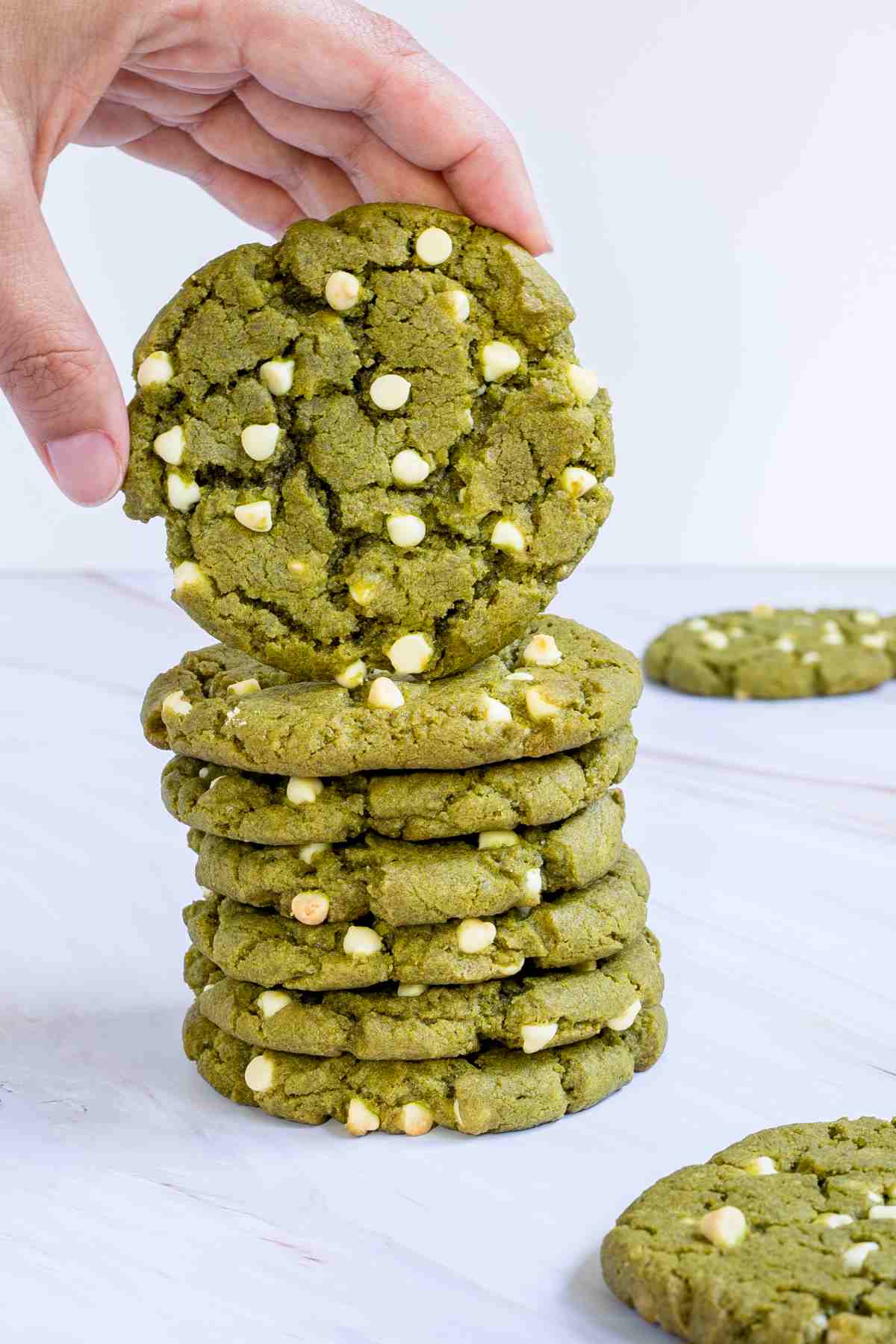 A stack of vibrant green cookies with white chocolate chips on a marble surface. A hand is taking one cookie from the top.