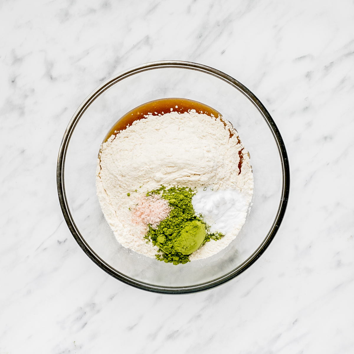A glass bowl with brown liquid, flour, and green matcha powder.