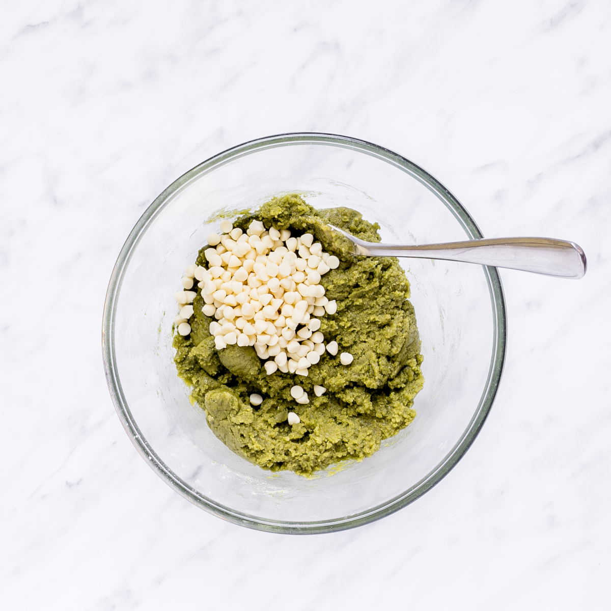 A glass bowl with green cookie batter and a heap of white chocolate chips.