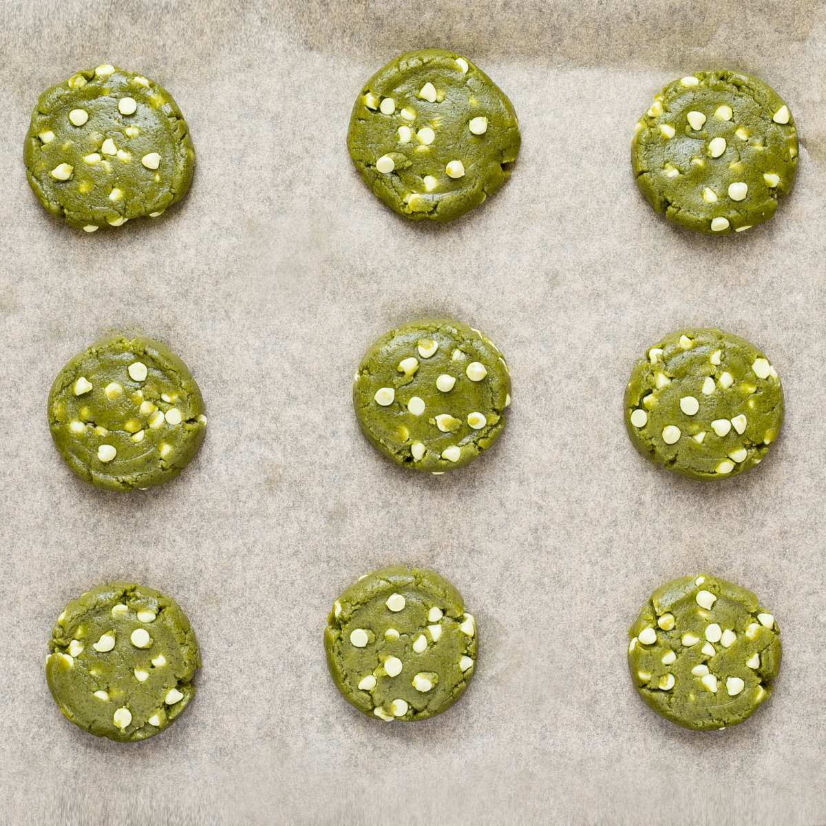 Unbaked round vibrant green cookie batters with white chocolate chips on parchment paper neatly placed away from each other.