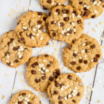 Lots of cookies with oats and chocolate chips on a wooden surface. More oats are sprinkled around them.