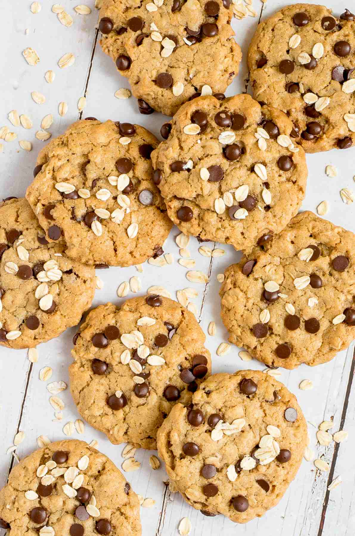 Lots of cookies with oats and chocolate chips on a wooden surface. More oats are sprinkled around them.