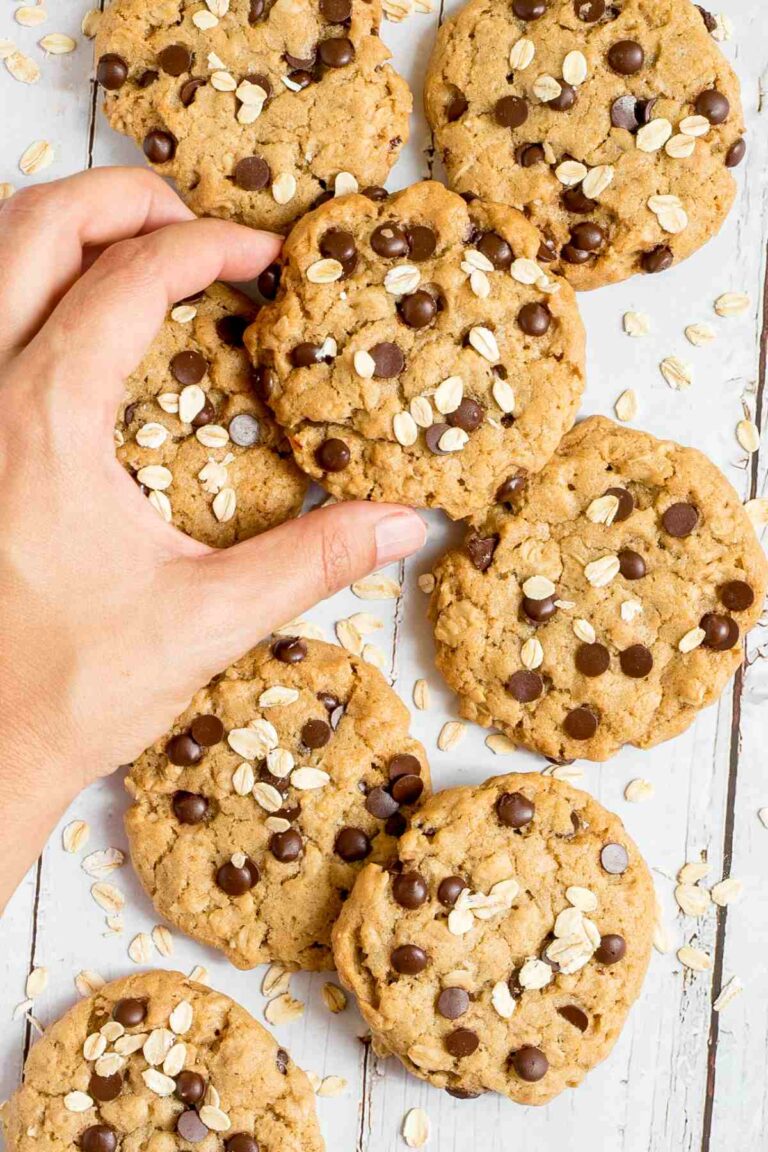 Lots of cookies with oats and chocolate chips on a wooden surface. More oats are sprinkled around them. A hand is taking one cookie for the middle.