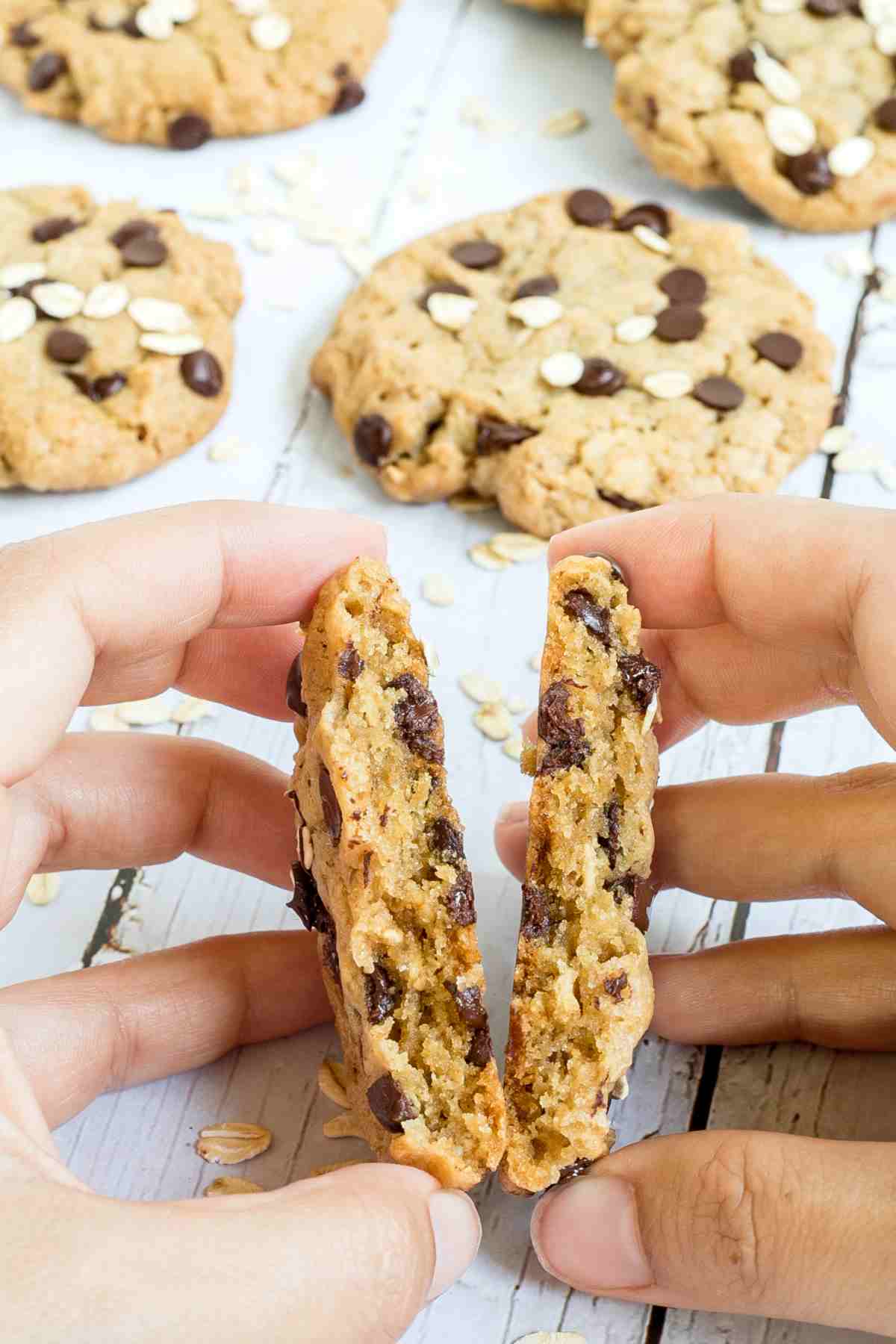 A hand is breaking one oatmeal chocolate chip cookie in half to show the texture. More cookies are in the background.