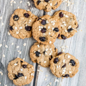 Lots of cookies with oats and raisins on a wooden surface. More oats are sprinkled around them.
