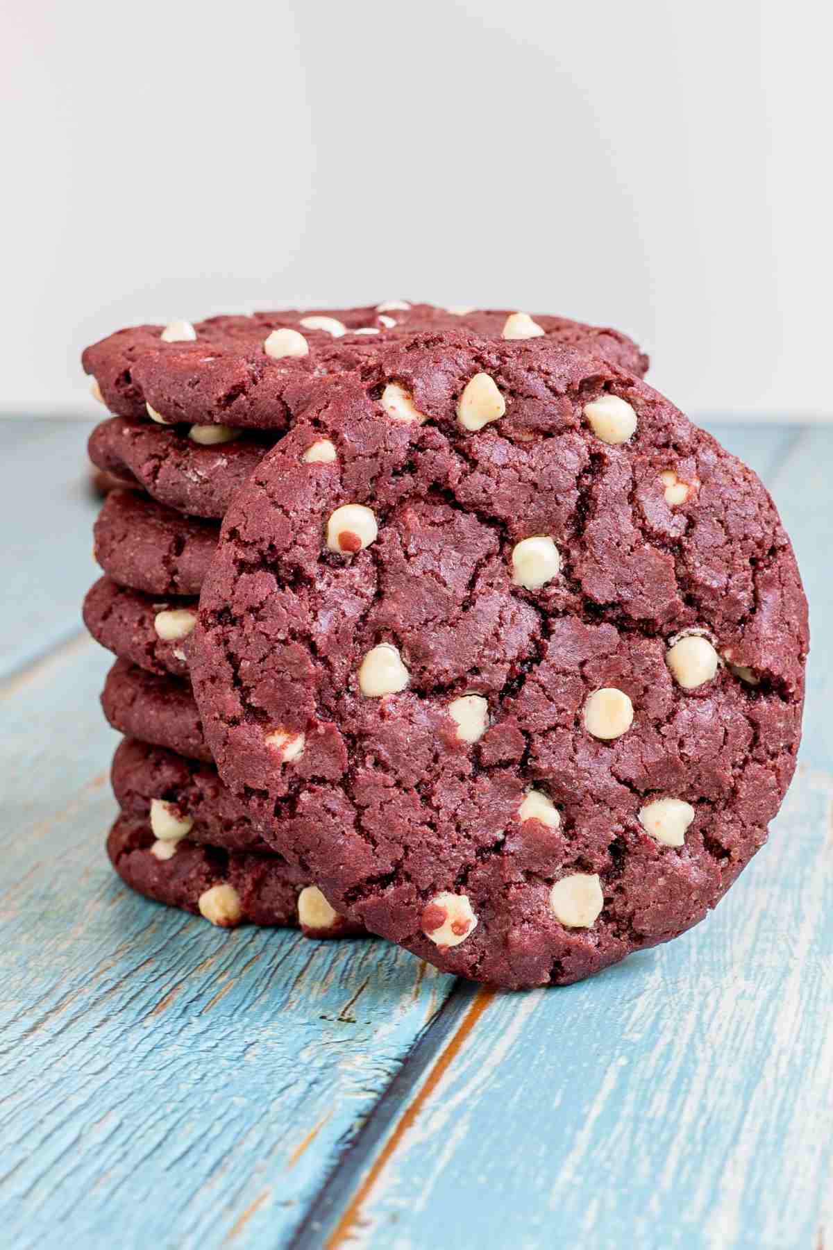 A stack red velvet cookies with white chocolate chips on a light blue wooden surface. One cookie is leaning to the side.