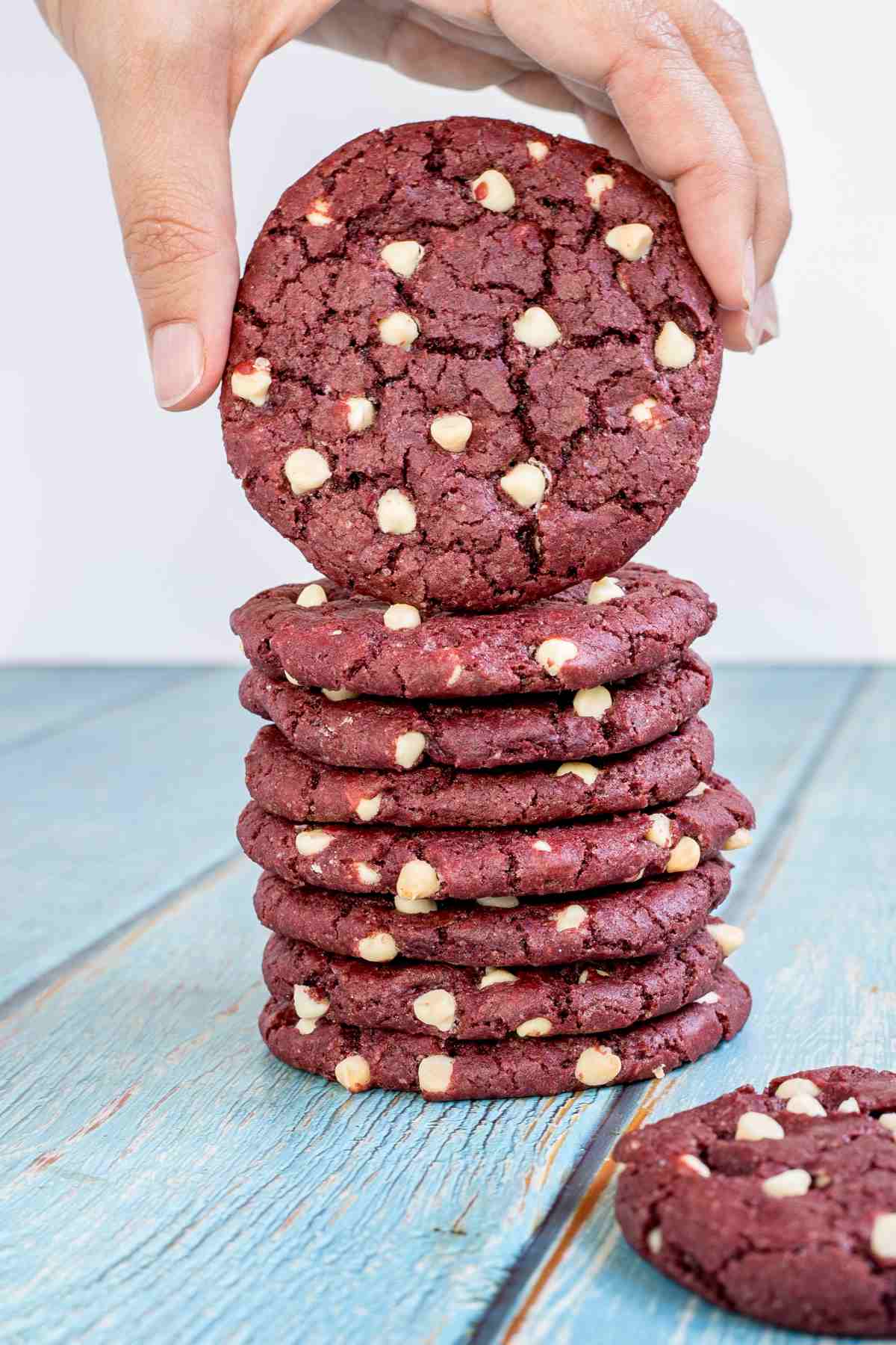 A stack of red velvet cookies with white chocolate chips on a light blue wooden surface. A hand is taking the top cookie.