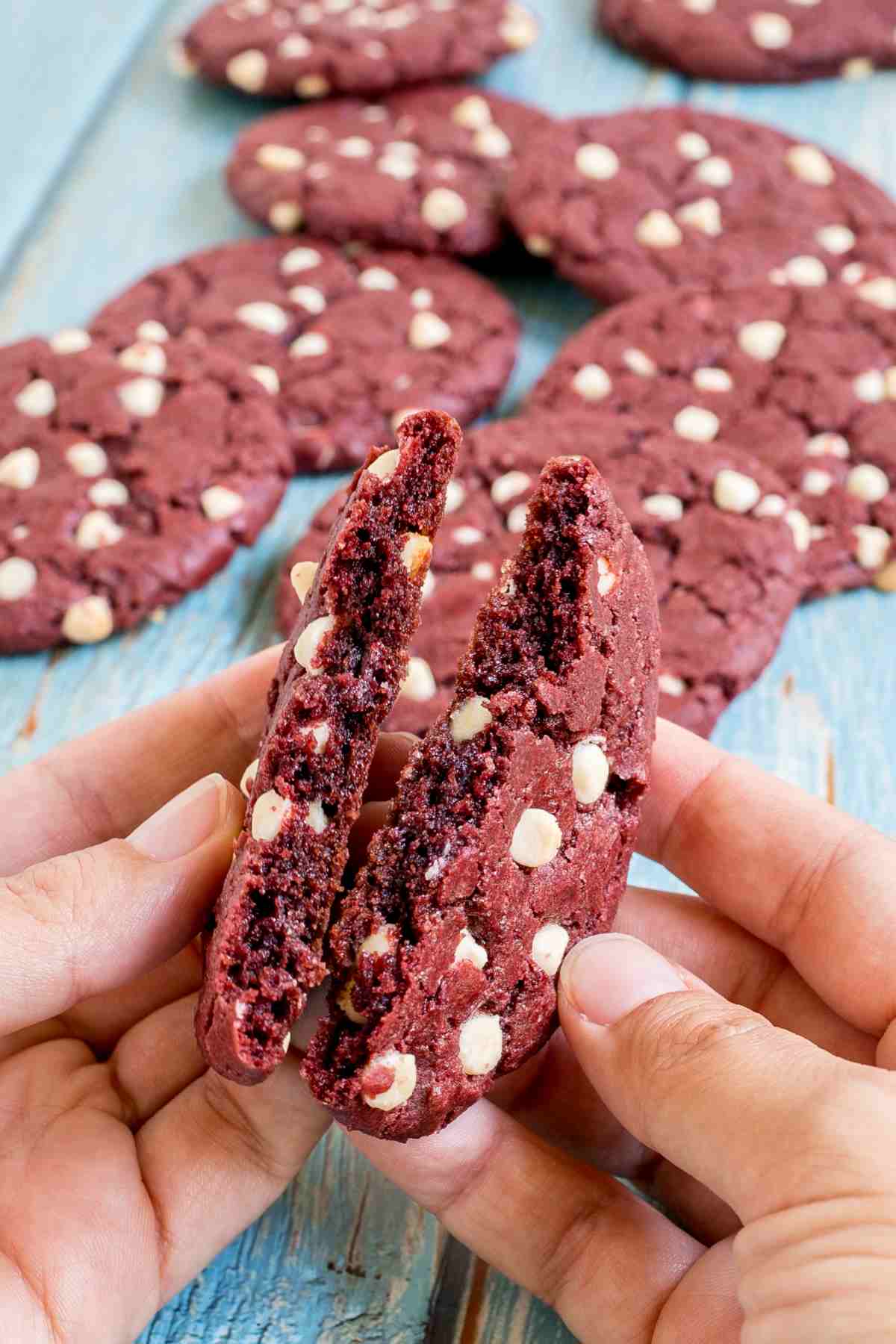 A hand is breaking a red velvet cookie with white chocolate chips in half and showing the texture. More cookies are in the background.