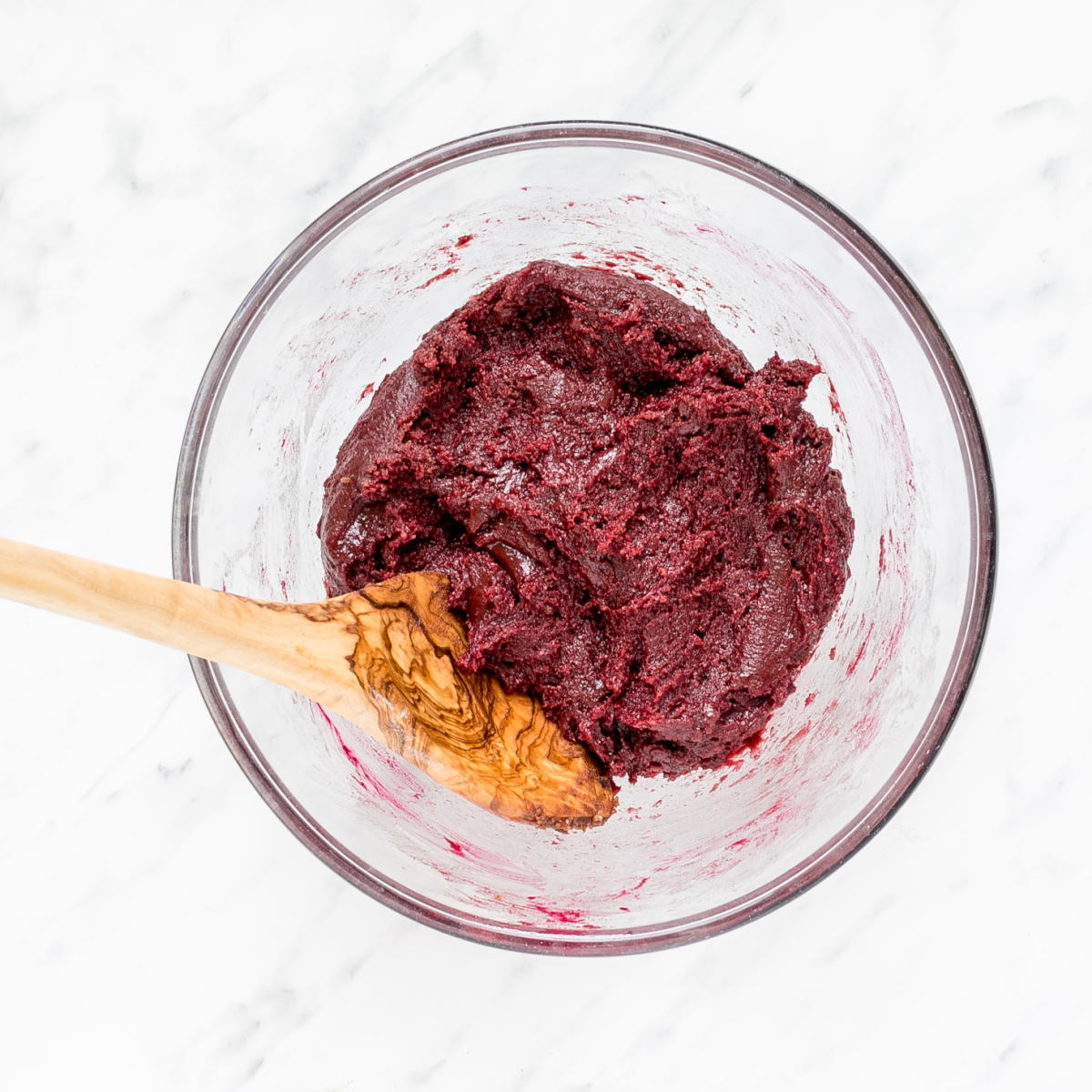 A glass bowl with red cookie dough and a wooden spatula.