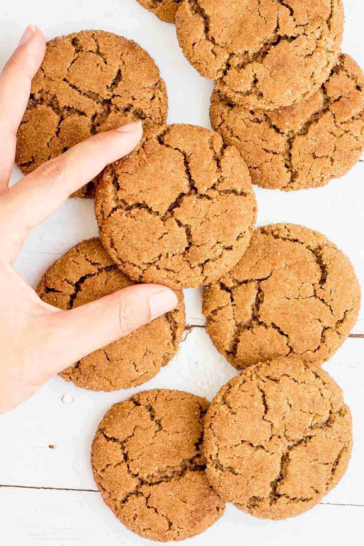 Light brown cookies with crinkled top on a white wooden surface. A hand is taking one from the middle.