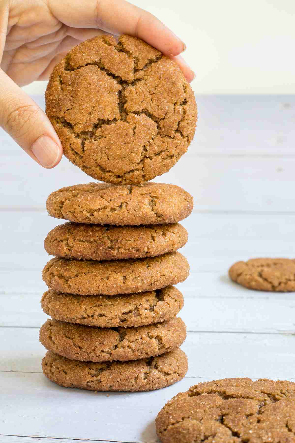 A stack of light brown cookies with crinkled top on a white wooden surface. A hand is taking one from the top.