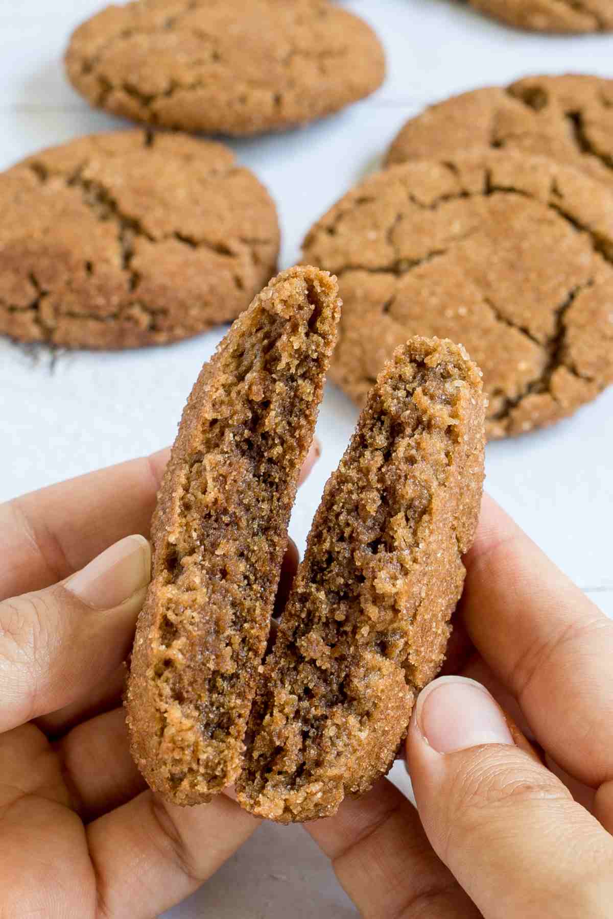 A hand is breaking a light brown cookie with a crinkled top in half to show the inner texture. More cookies are behind it.