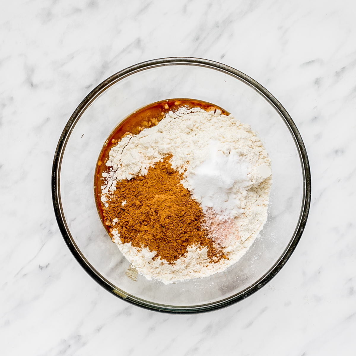 A glass bowl with brown liquid and heaps of flour, cinnamon, baking powder, and salt.