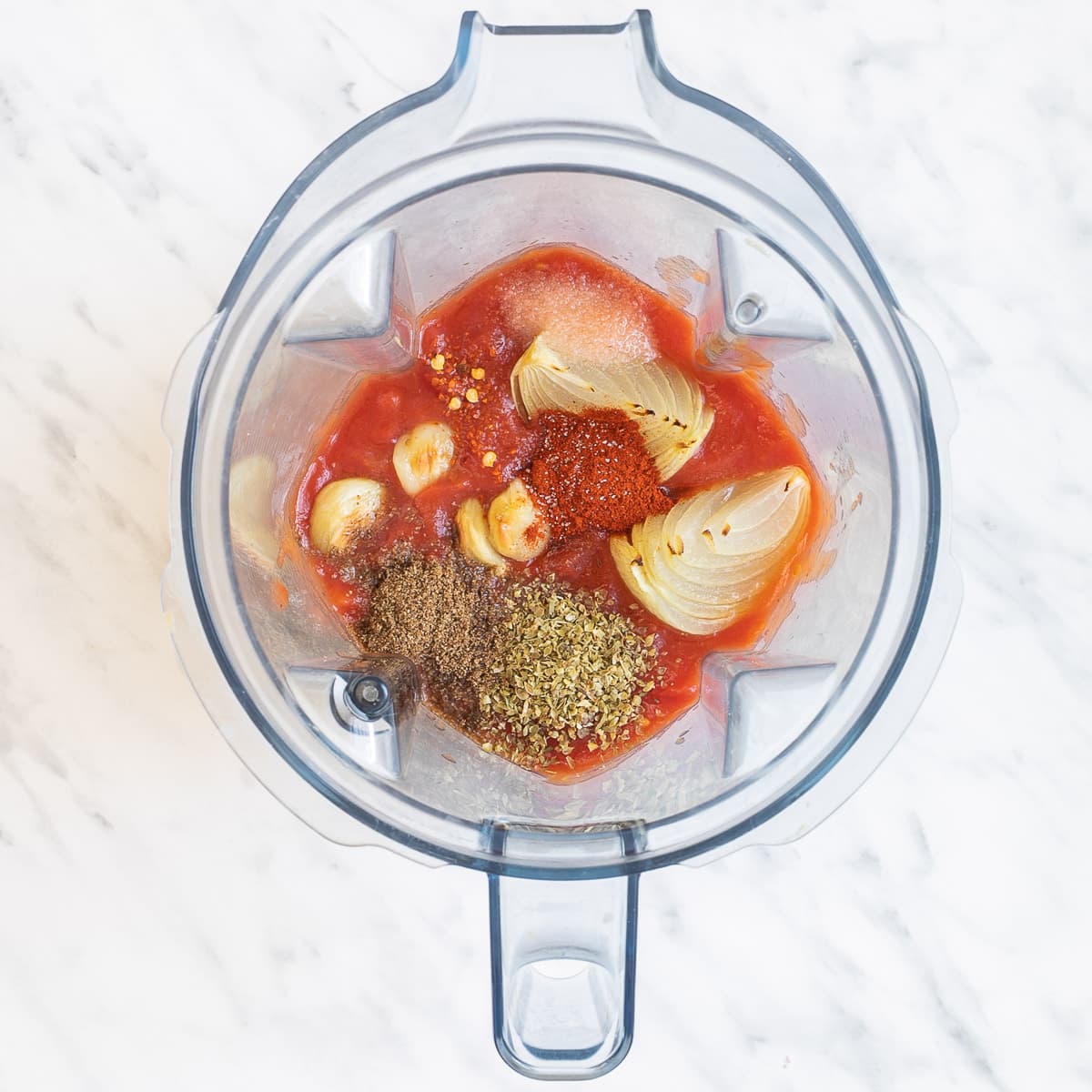 A blender with red sauce, garlic, onion, and small heaps of different spices and herbs.