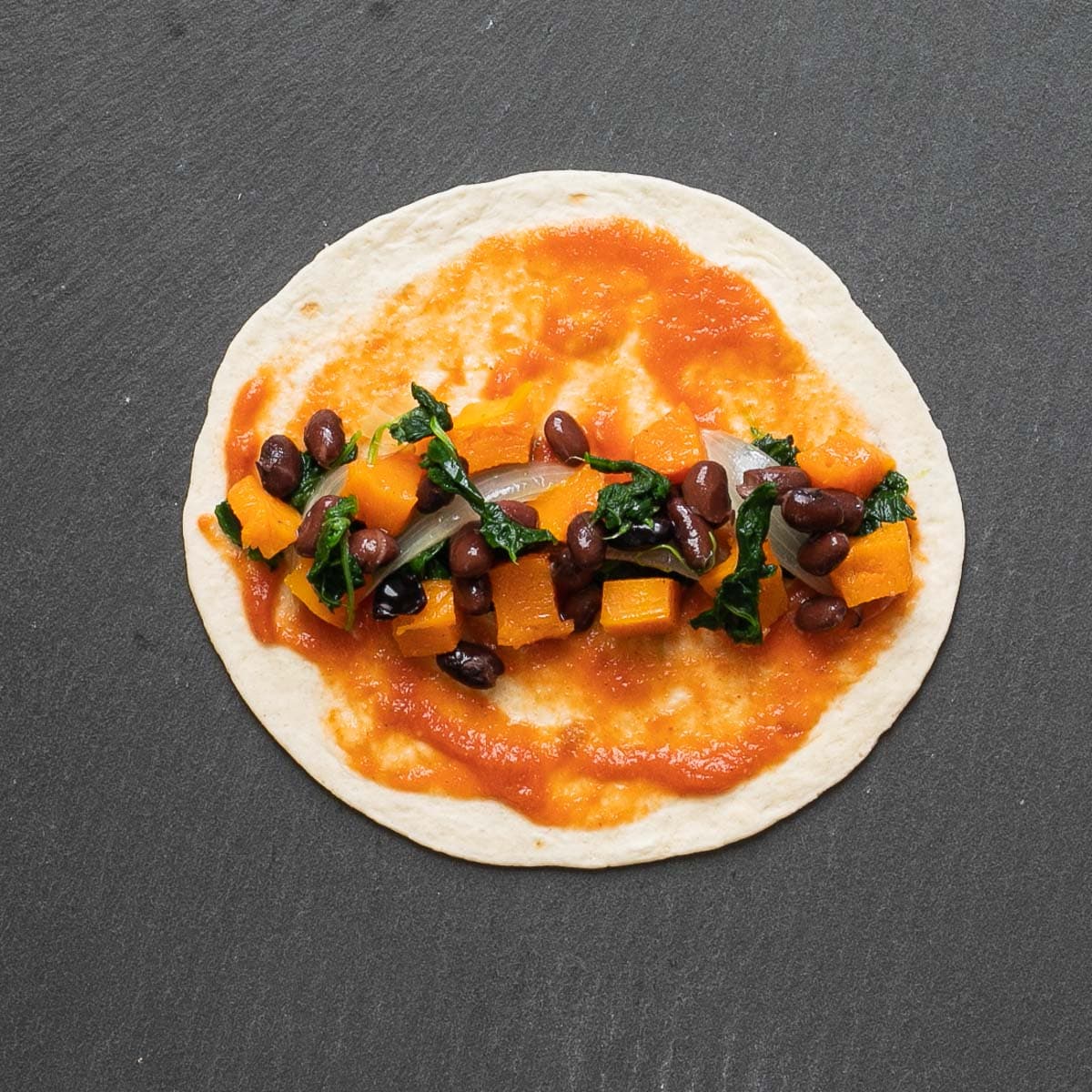 A small tortilla with red sauce spread on top and spinach leaves, black beans, and orange squash cubes in the middle.