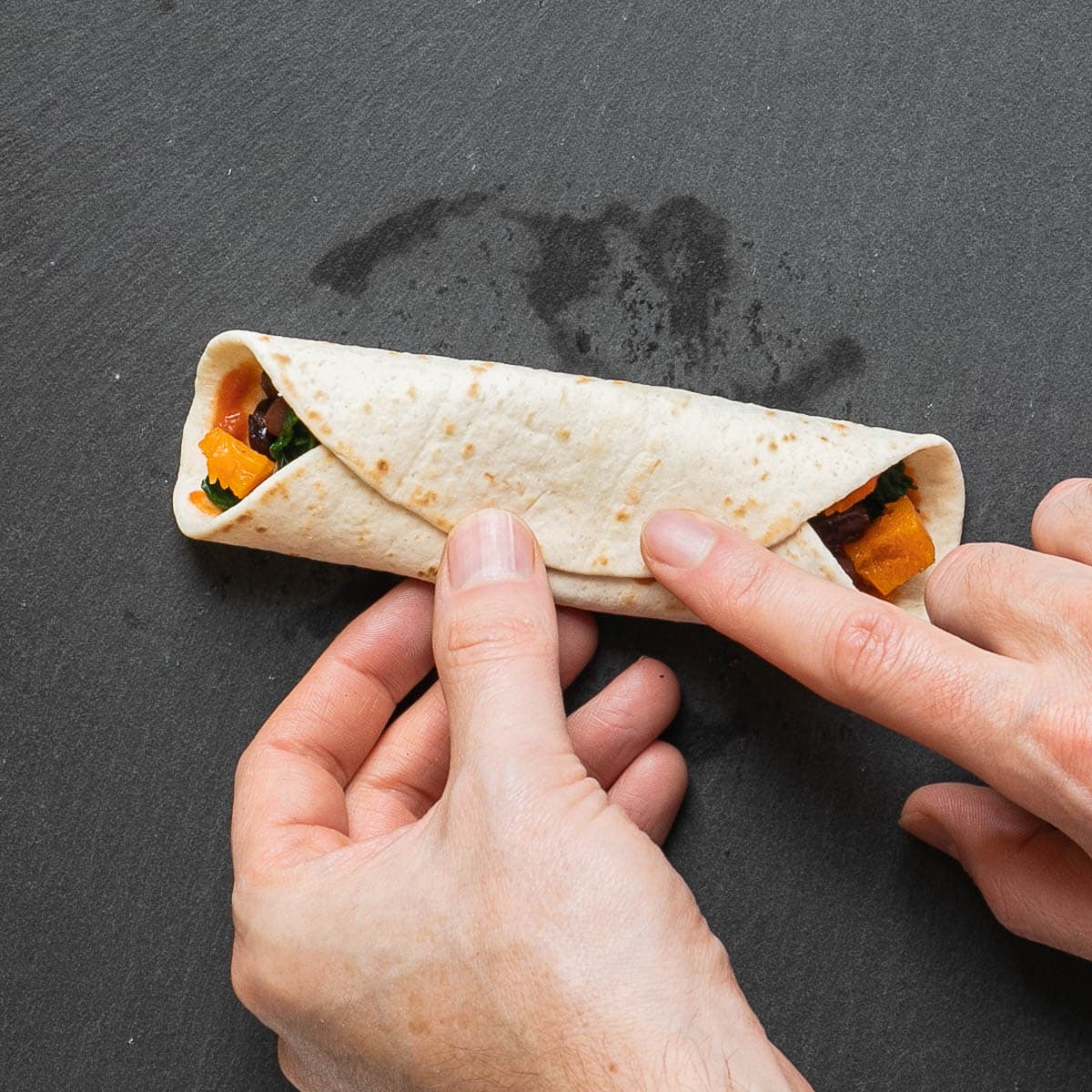 A hand is rolling a small tortilla filled with red sauce, black beans and butternut squash cubes.
