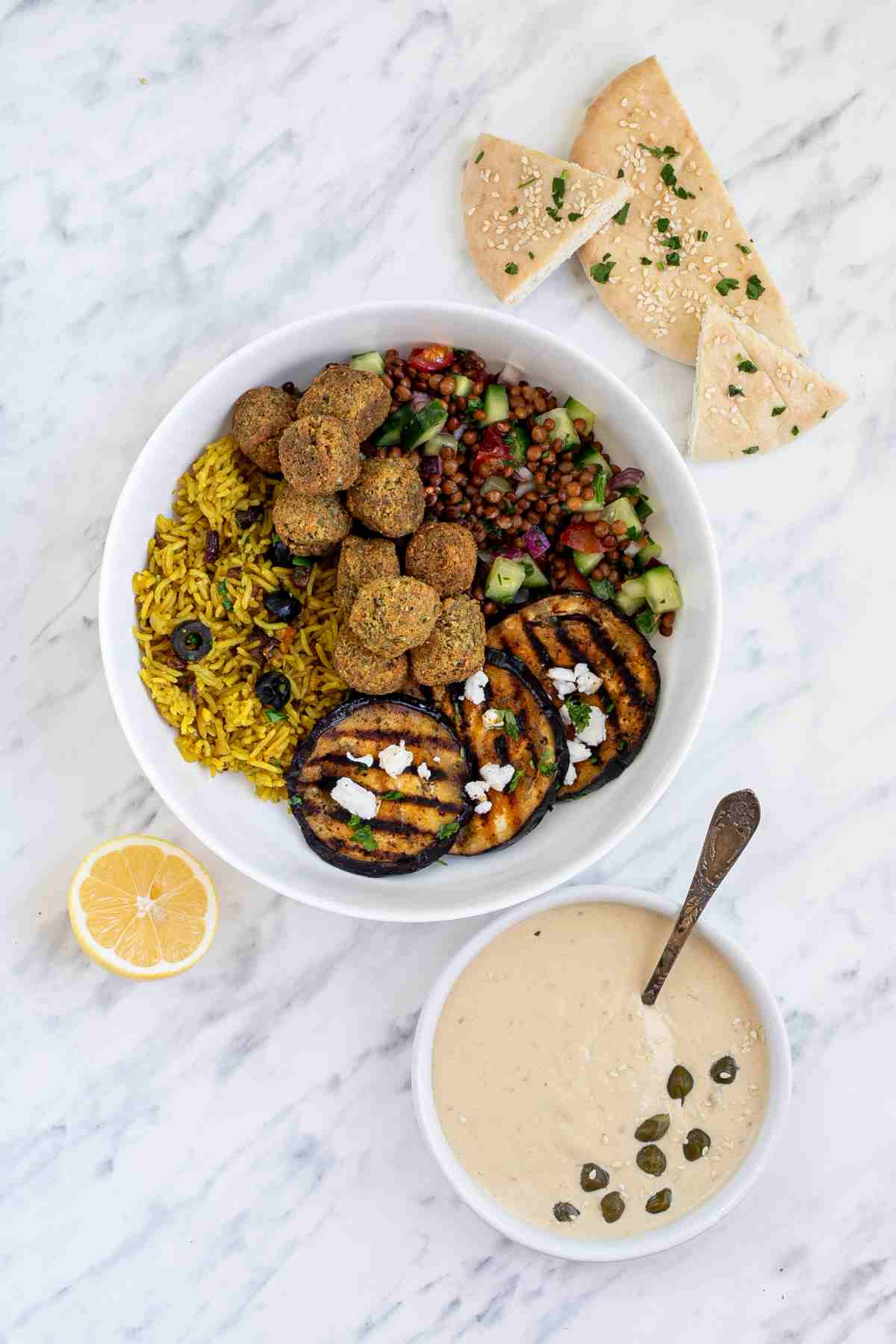 A large white bowl with falafel balls, grilled eggplant, yellow rice and chopped tomato and cucumber salad with lentils. Light brown sauce is next to it as well as half lemon and naan bread.