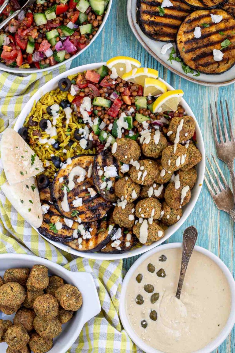 A large white bowl with falafel balls, grilled eggplant, yellow rice and chopped tomato and cucumber salad with lentils drizzled with a white sauce. The remaining ingredients are in bowls around the main one.