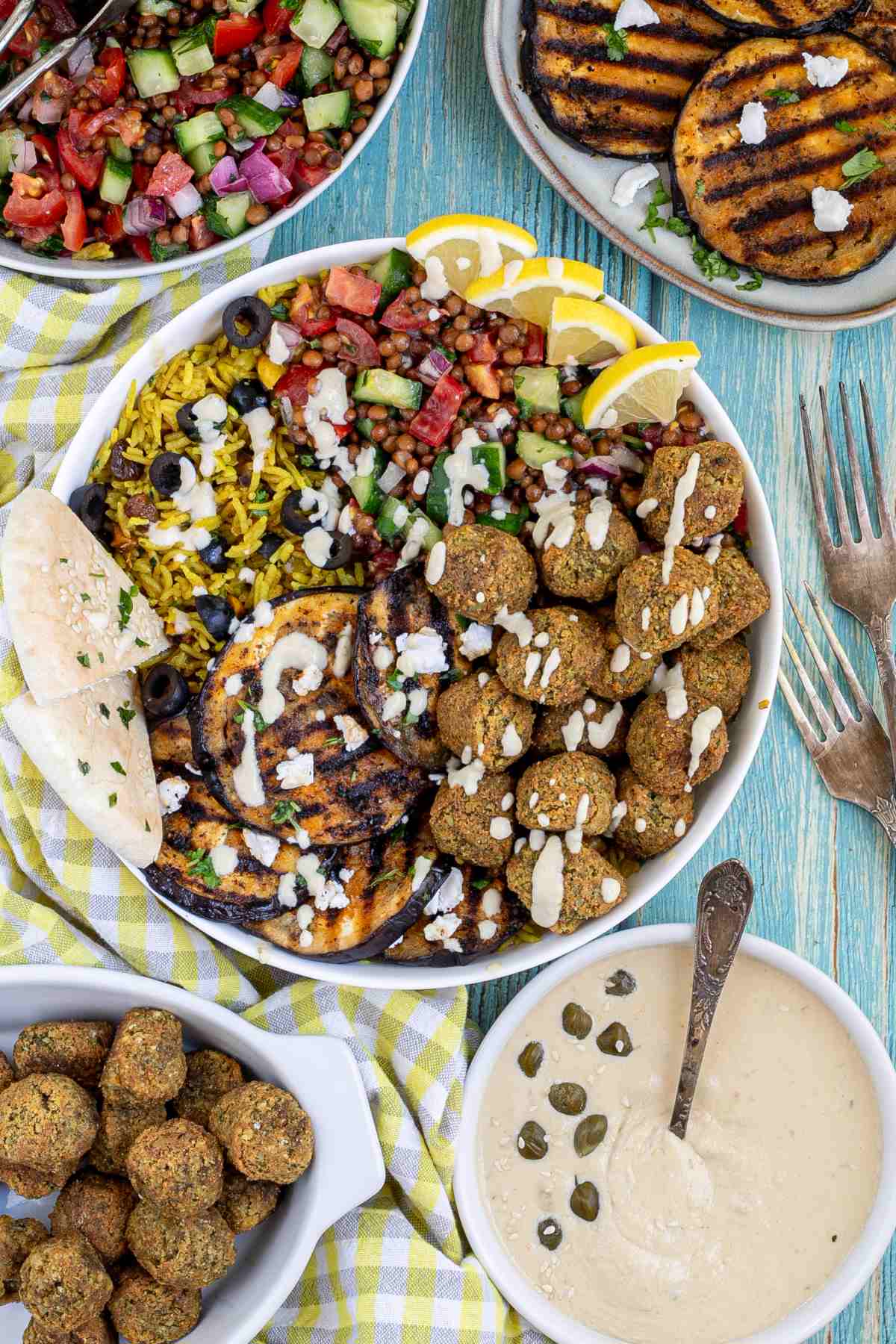A large white bowl with falafel balls, grilled eggplant, yellow rice and chopped tomato and cucumber salad with lentils drizzled with a white sauce. The remaining ingredients are in bowls around the main one. 