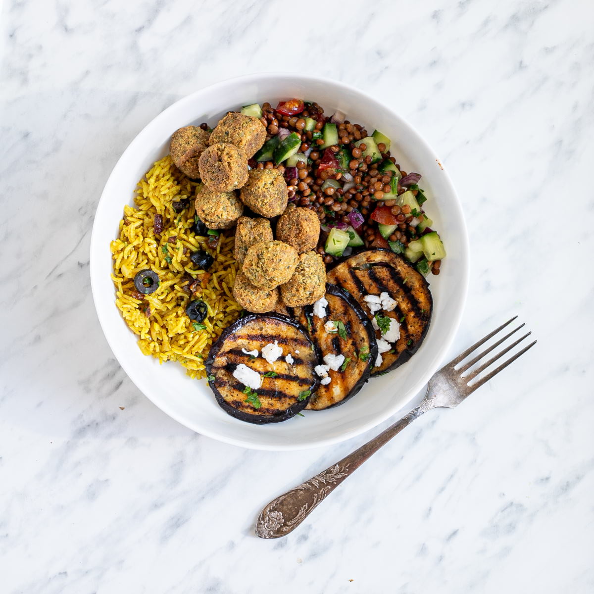 A large white bowl with falafel balls, grilled eggplant, yellow rice and chopped tomato and cucumber salad with lentils.